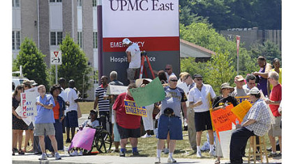 Protesters demonstrate in front of the new UPMC East facility in Monroeville in July, as workers put the finishing touches on the sign.